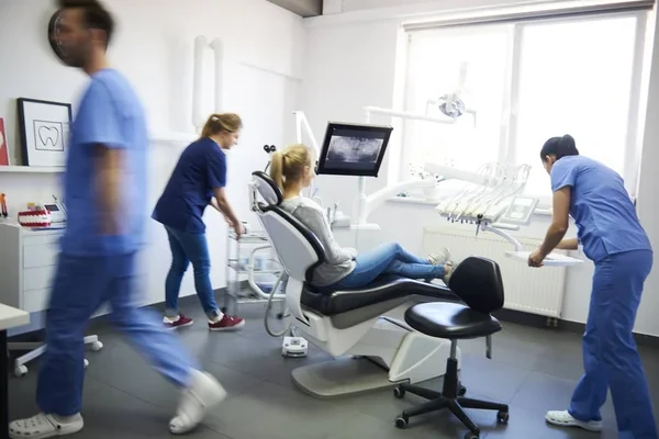 A busy, modern dental clinic room with a patient in the chair and staff preparing for a dental procedure