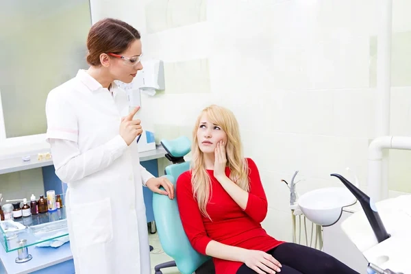A dental patient holding her cheek in pain while describing severe toothache symptoms to a dentist during a consultation