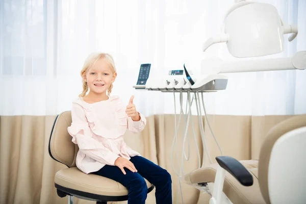 A happy young girl giving a thumbs-up while sitting in a pediatric dentist's office