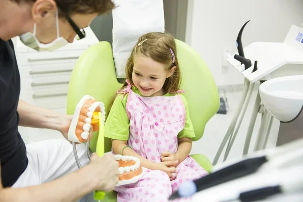 A pediatric dentist using an oversized tooth model and toothbrush to demonstrate brushing techniques to a smiling young girl.