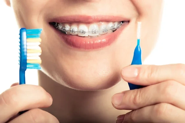 A smiling woman wearing metal dental braces holding a standard toothbrush and an interdental brush for orthodontic cleaning