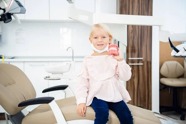A smiling young girl holding a dental jaw model while sitting comfortably in a pediatric dentist's chair.