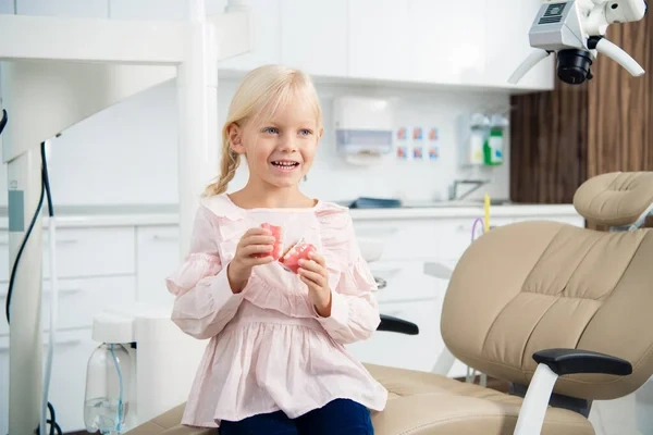 A smiling young girl holding a two-piece dental model while sitting in a pediatric dentist's chair.