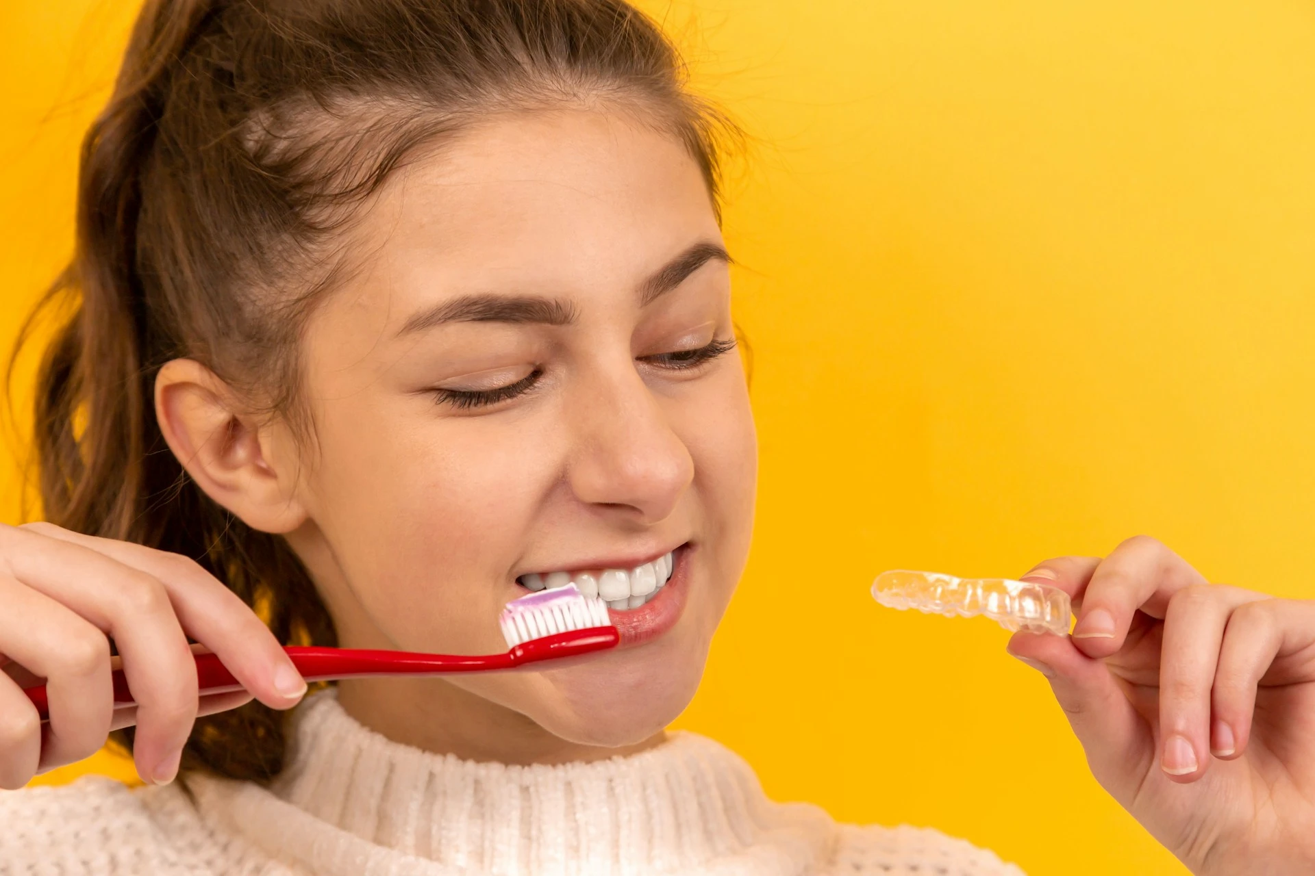 A young woman brushing her teeth while holding a clear Invisalign aligner tray