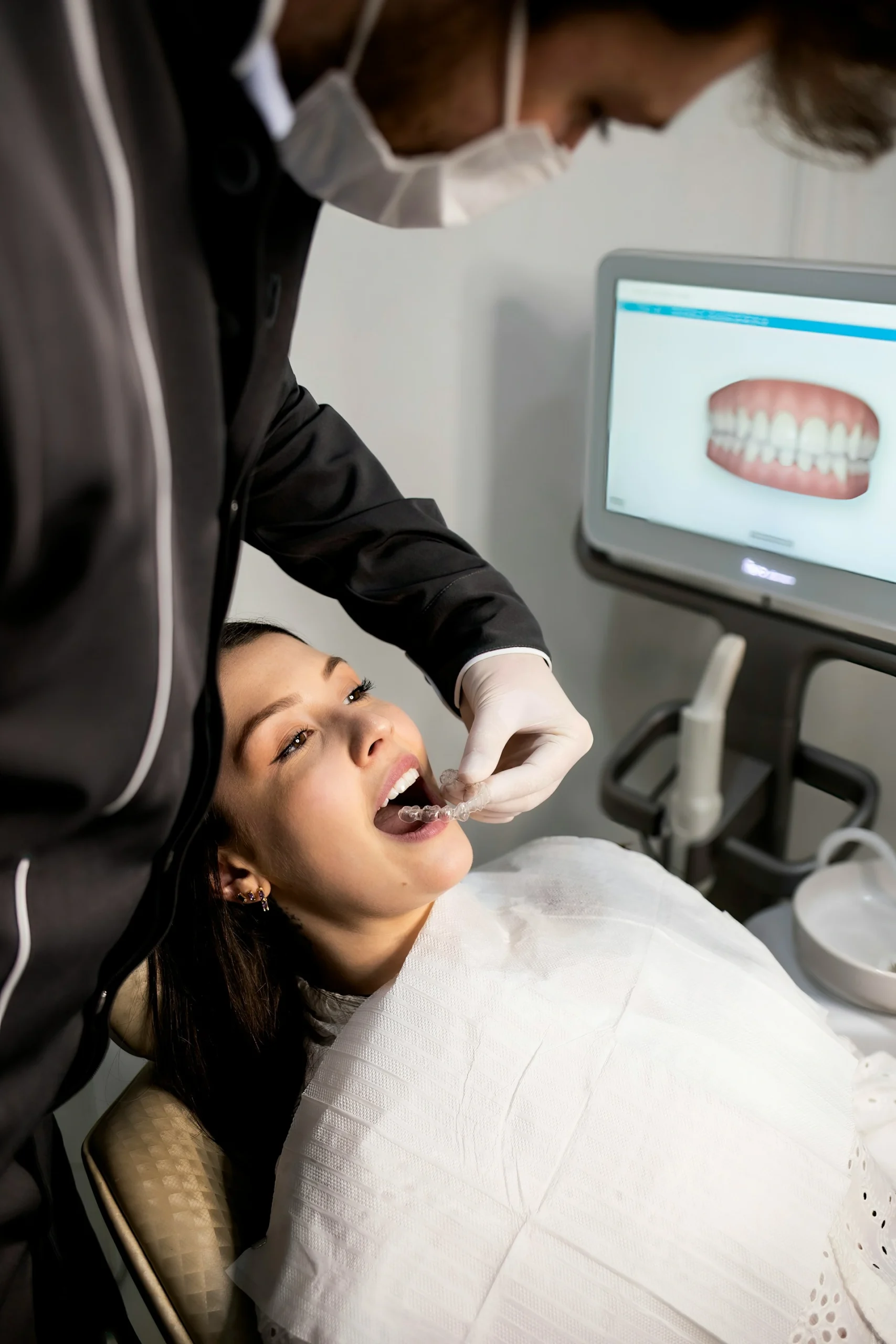 A dentist wearing a black uniform, face mask, and white gloves placing a clear plastic aligner tray into a female patient's mouth, with a 3D dental scan model displayed on a computer monitor screen in the background.