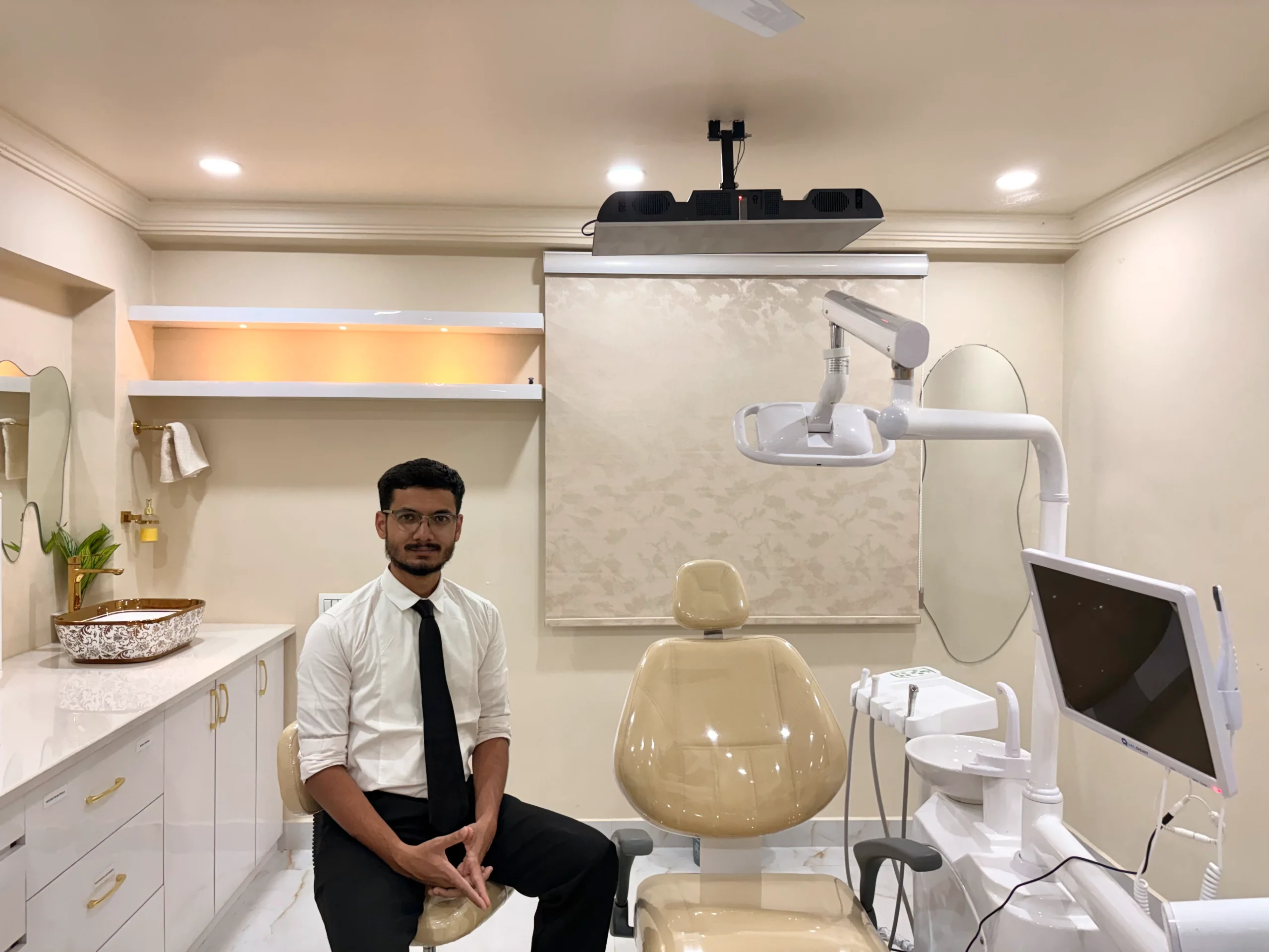 Dr. Ahmed in formal attire sitting by the patient chair inside a clean treatment room at Unique Dental.